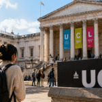 view of University College London campus on a sunny day, with students walking near the historic main building and a student carrying a backpack facing a large UCL sign